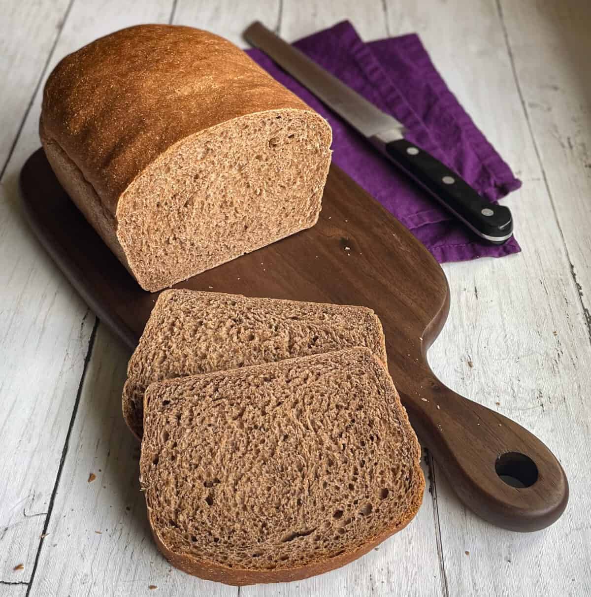 loaf of brown bread on a cutting board with two slices, knife, and napkin.