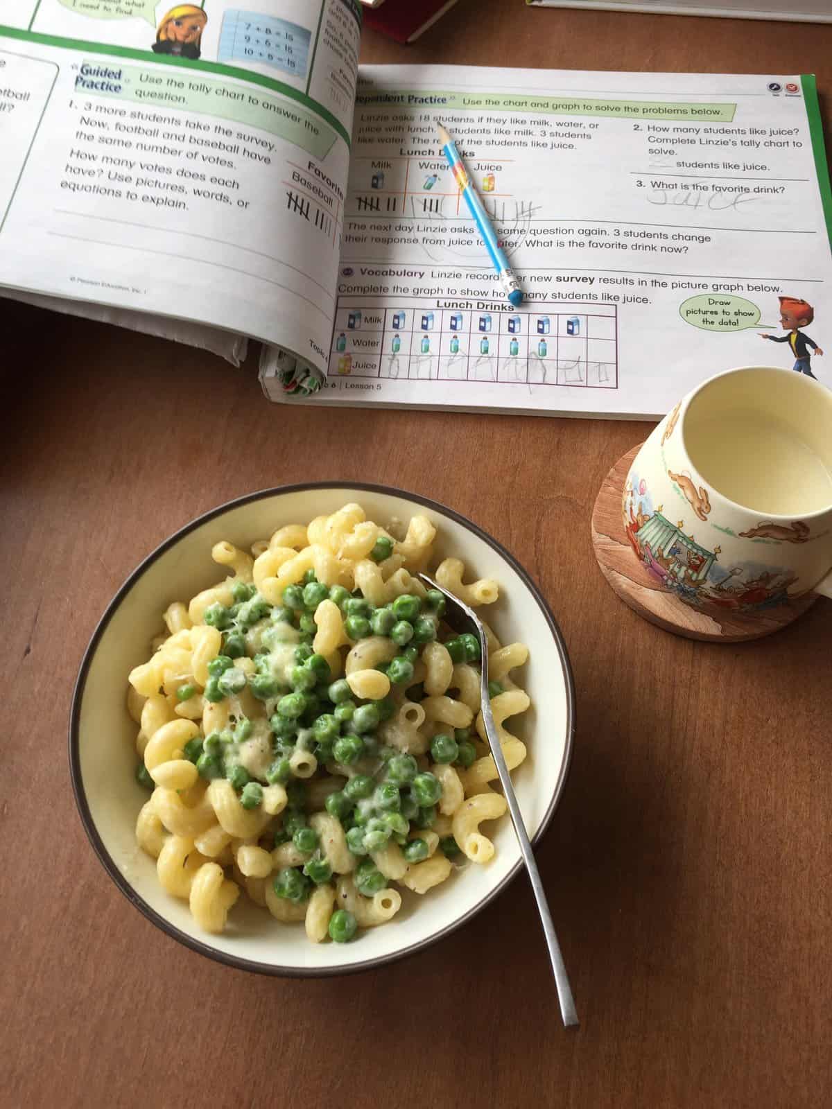 bowl of cavatappi alfredo with peas and Parmesan, cup of milk, and child's workbook.