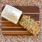 funfetti loaf cake with white glaze cut into slices on a cutting board surrounded by rainbow sprinkles.