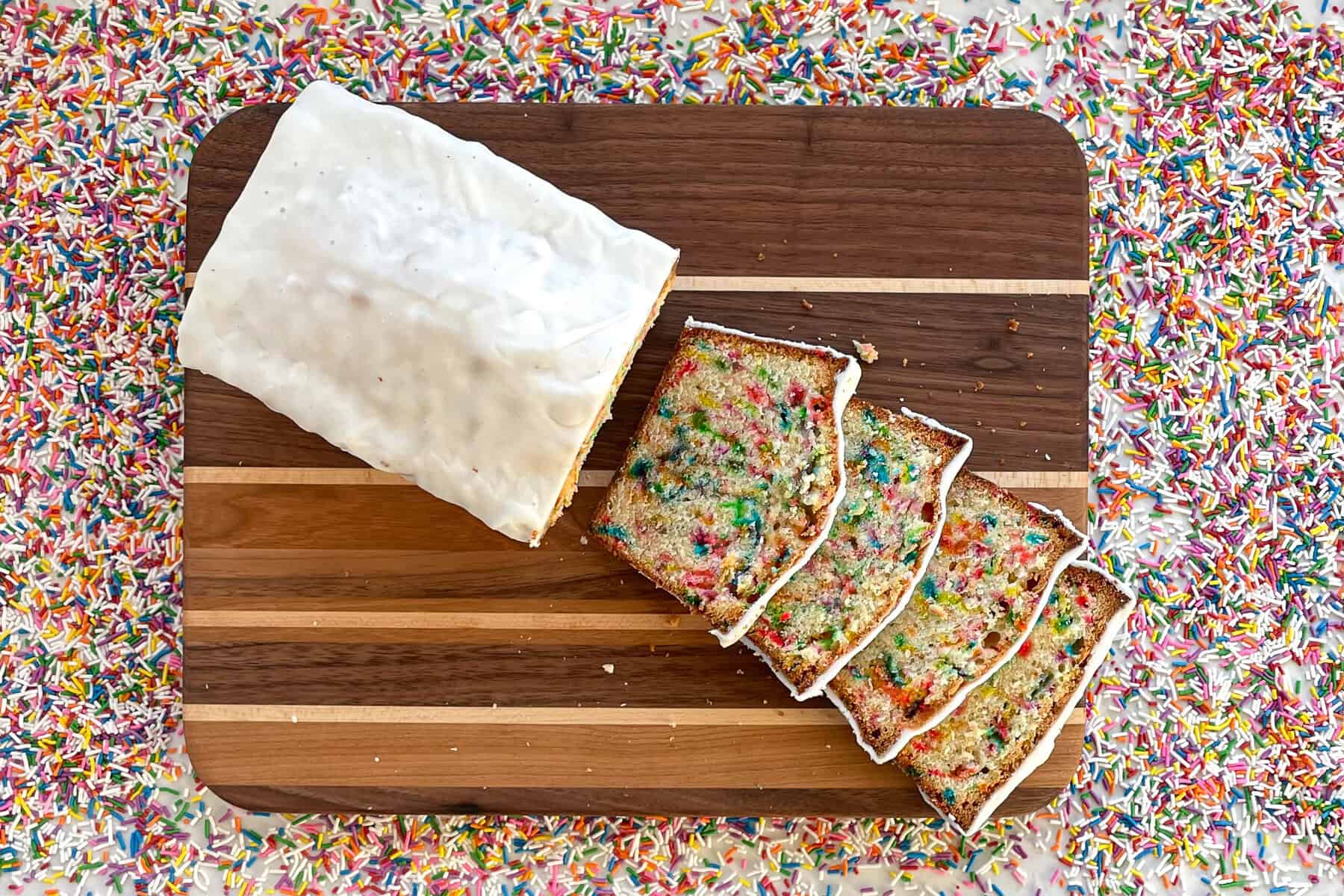 sliced funfetti loaf cake with white glaze on cutting board surrounded by rainbow sprinkles.