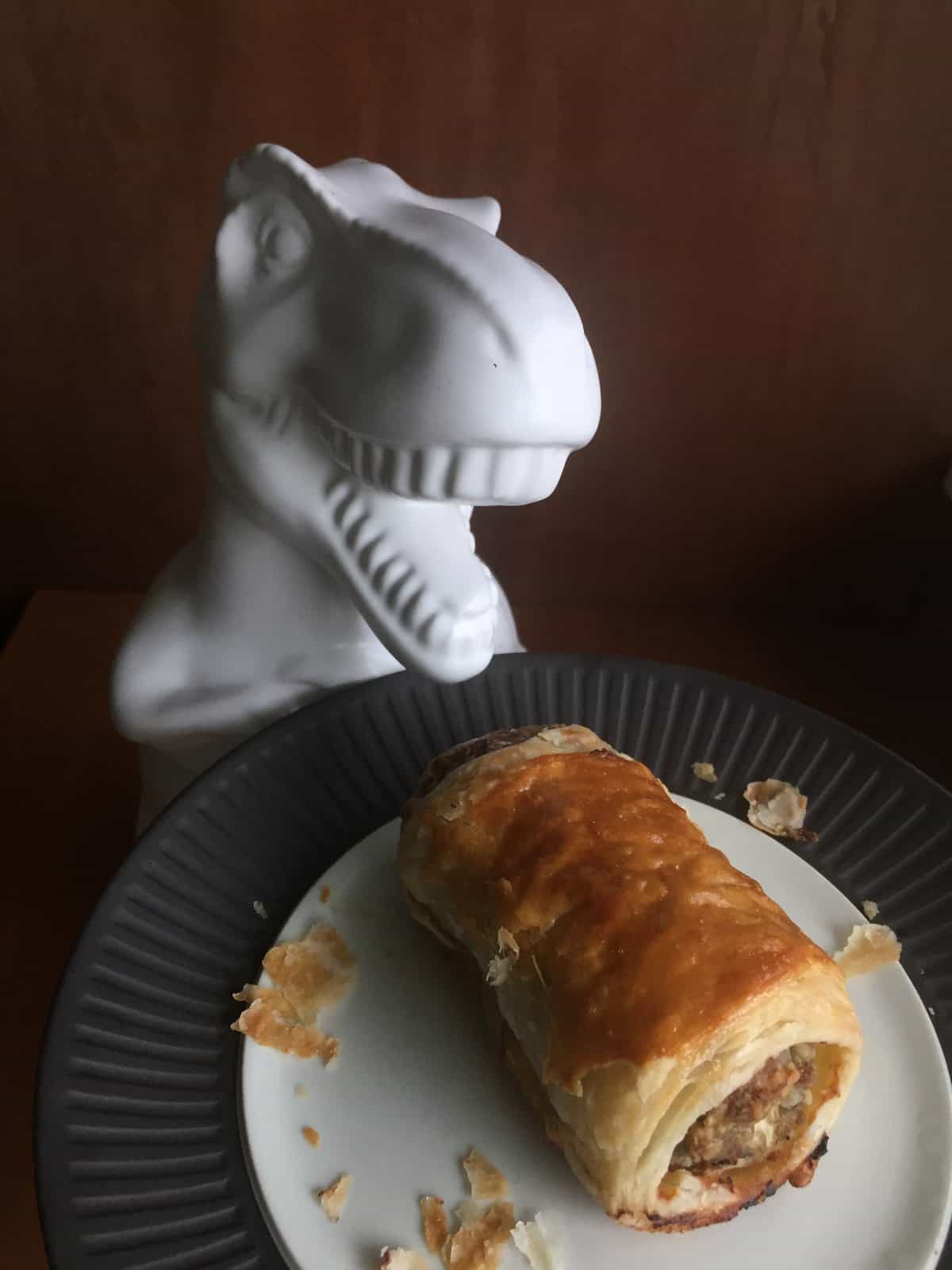a dinosaur cookie jar about to eat a puff pastry sausage roll on a plate.