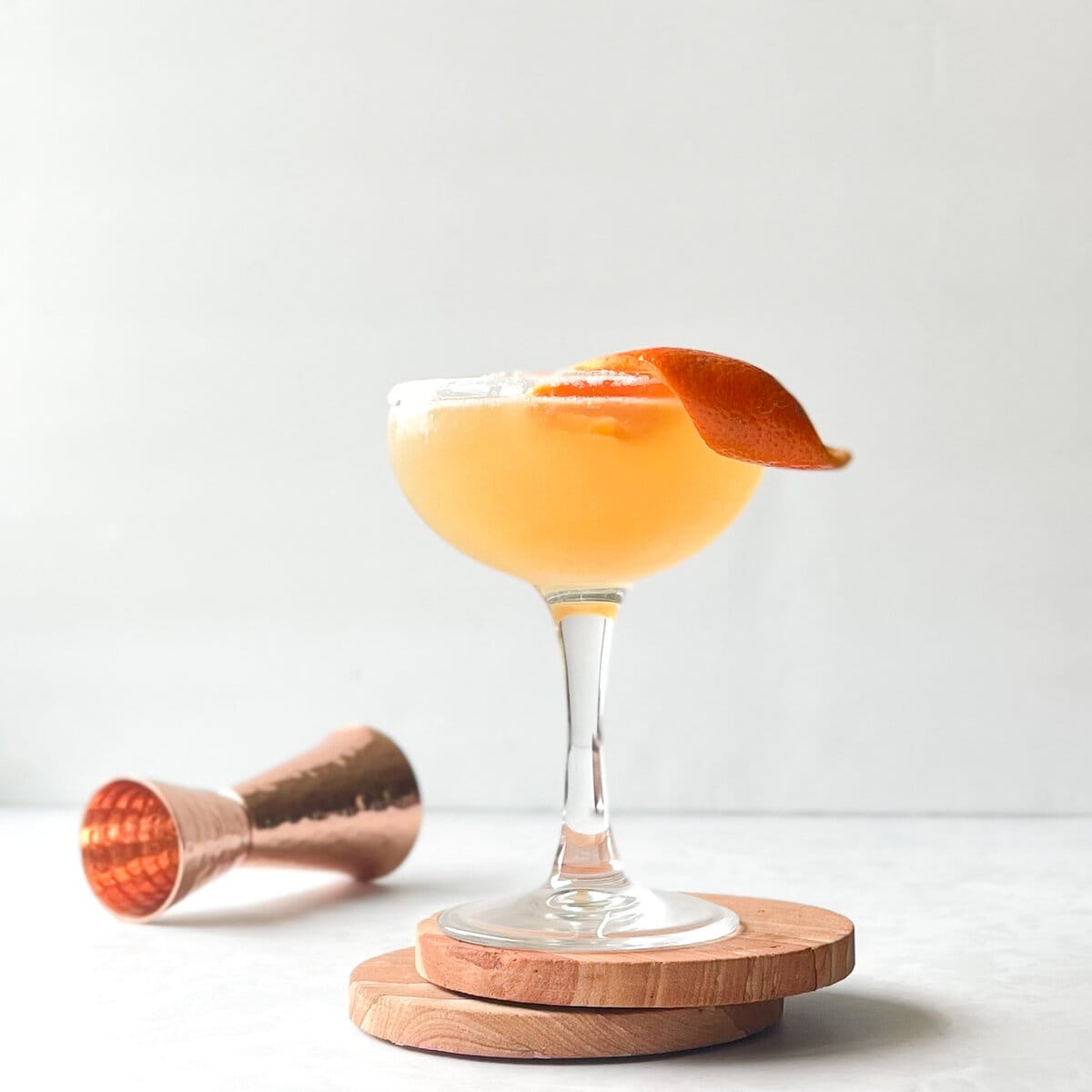 pale orange cocktail in a coupe garnished with orange twist, on two coasters and a jigger.