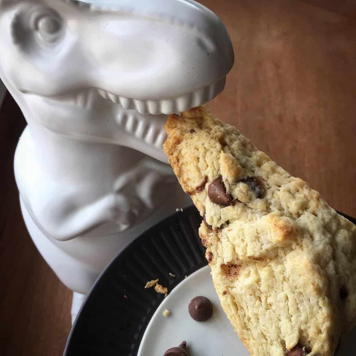 Tyranosaurus rex cookie jar taking a bite out of a chocolate chip scone on a plate.