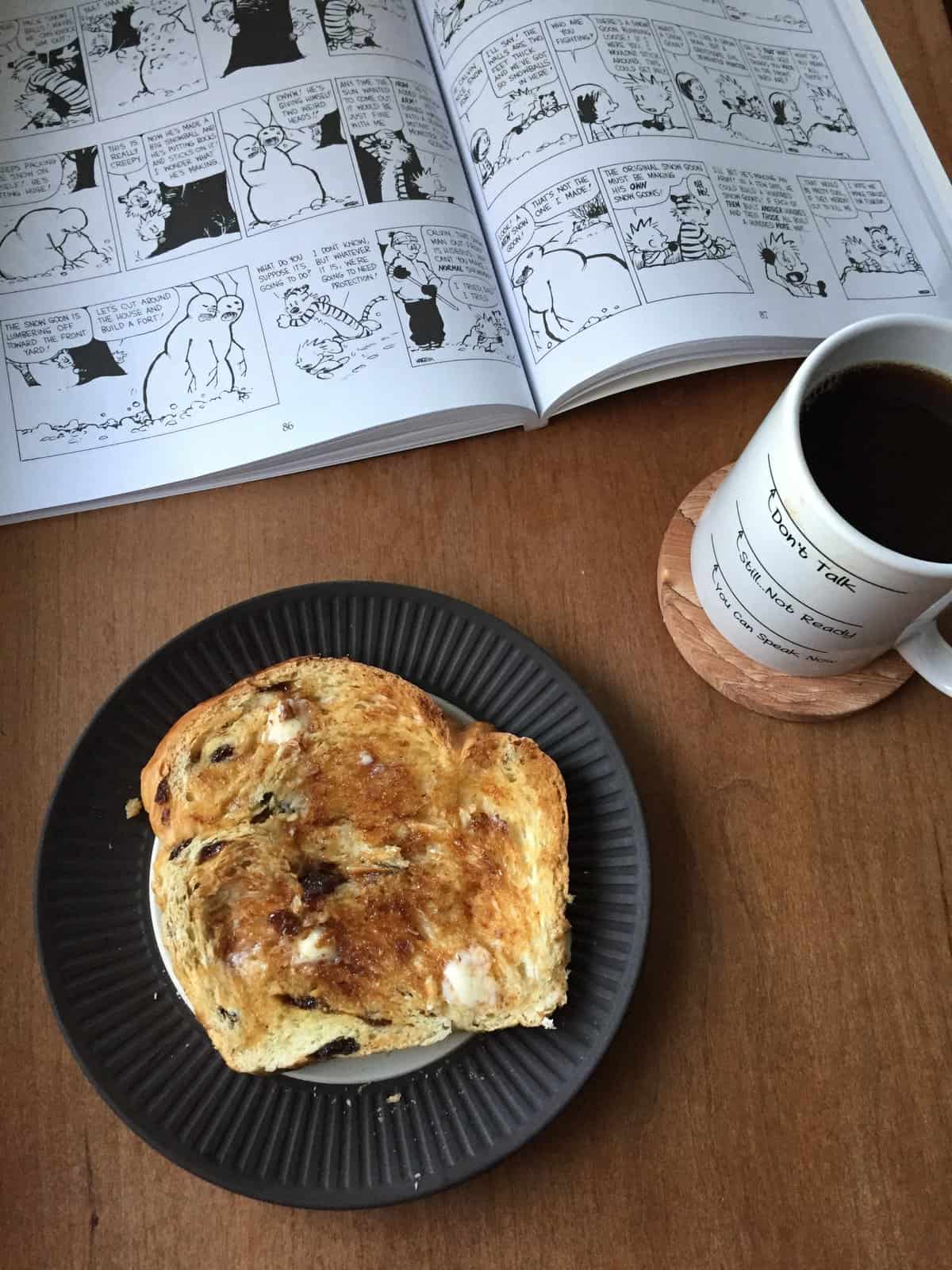 a slice of toasted raisin challah on a plate with coffe cup and comic book.