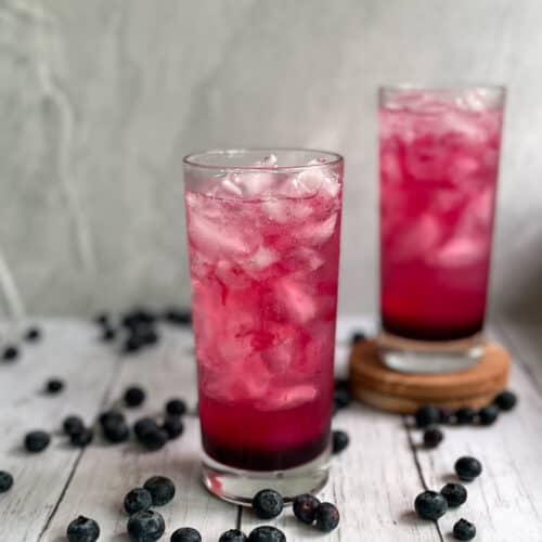 two deep pink blueberry gin and tonics surrounded by fresh blueberries.
