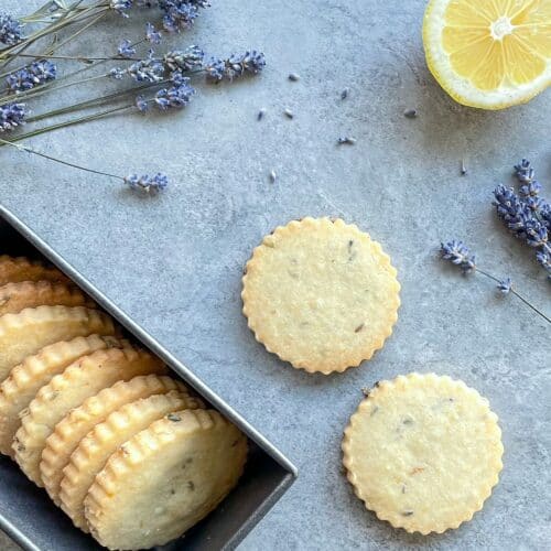 lavender flecked shortbread cookies nestled vertically in a metal sleeve next to two cookies with lavender blossoms and lemon half on a countertop.