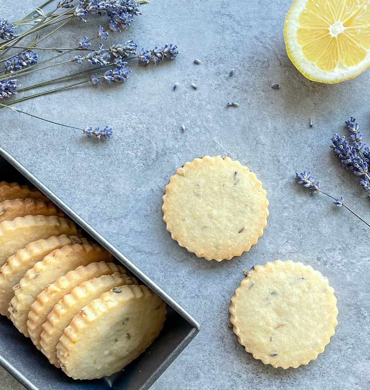 lavender flecked shortbread cookies nestled vertically in a metal sleeve next to two cookies with lavender blossoms and lemon half on a countertop.