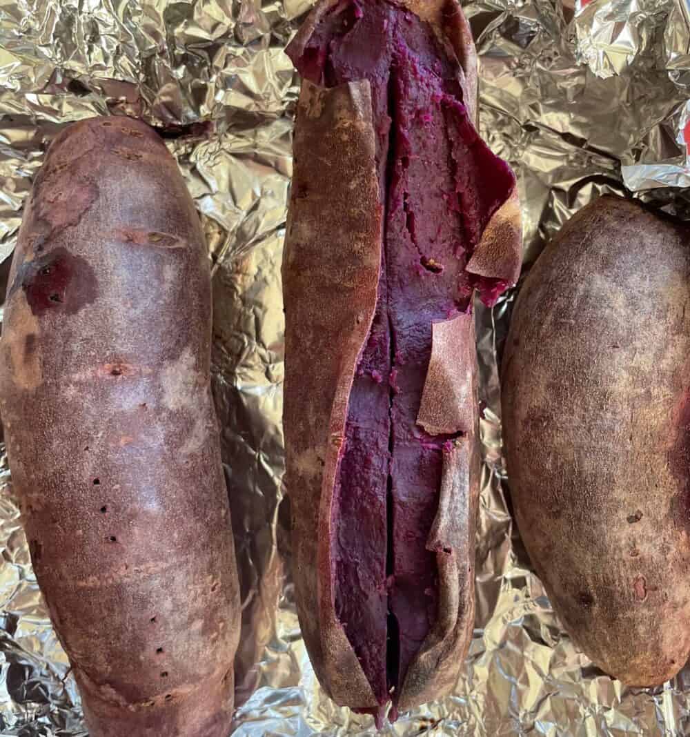 three purple sweet potatoes on foil with one sliced open to show the purple flesh.