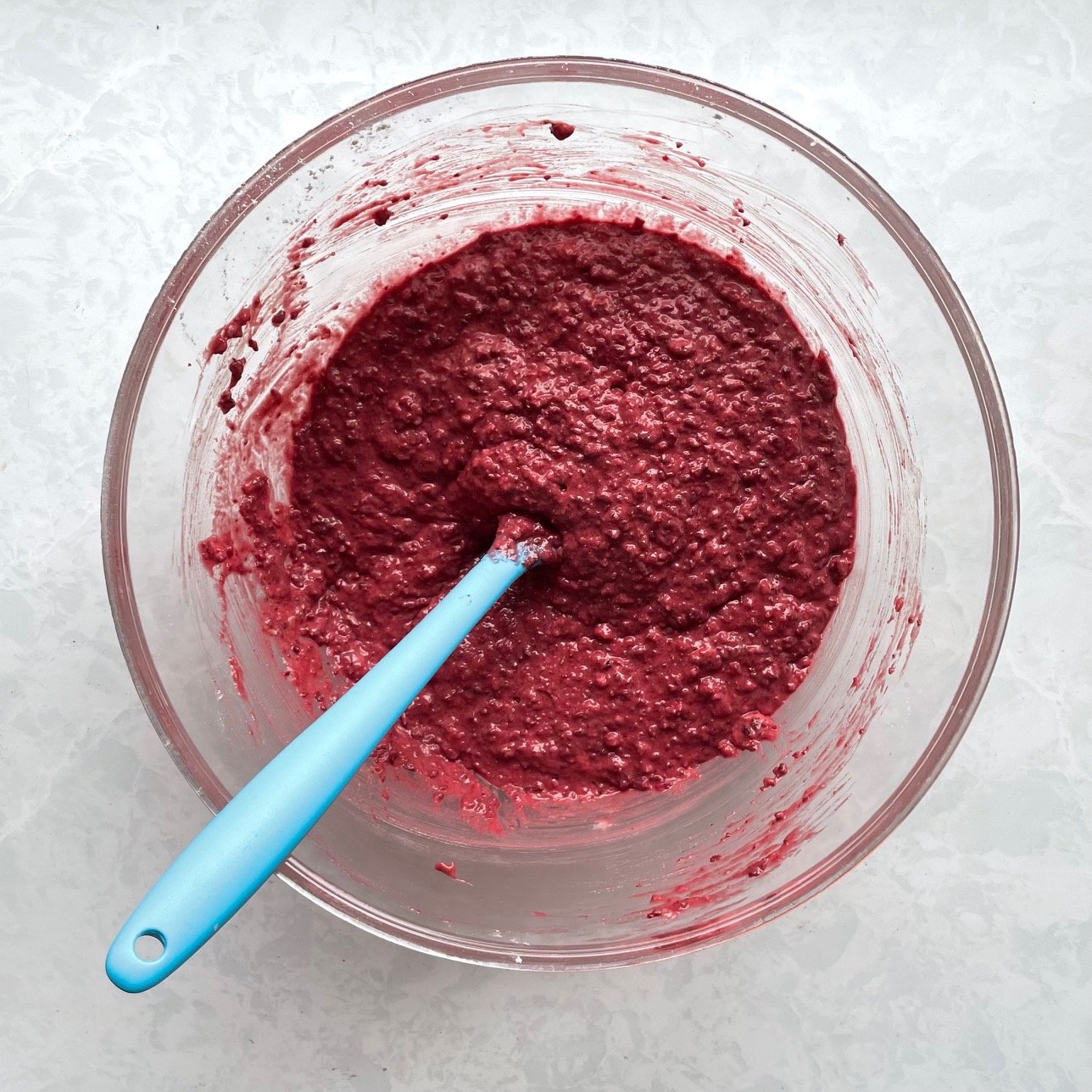 red waffle batter with a blue silicone spatula in a glass bowl.