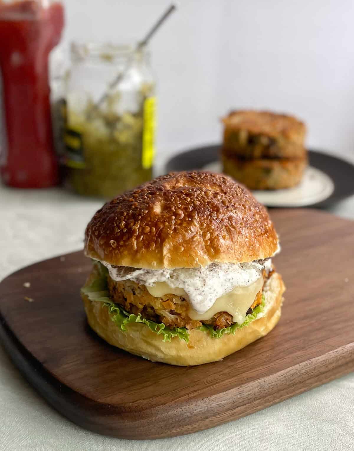 a cauliflower burger on a pretzel bun with cheese and lettuce with ketchup and relish in background.