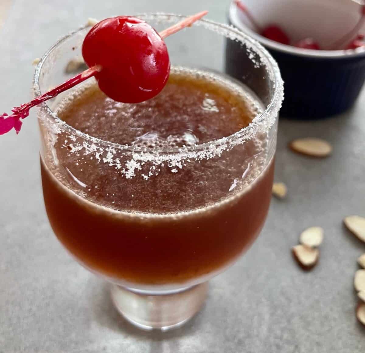 a cherry bakewell cocktail with a sugared rim and maraschino cherry, scattered almonds, and a bowl of cherries.