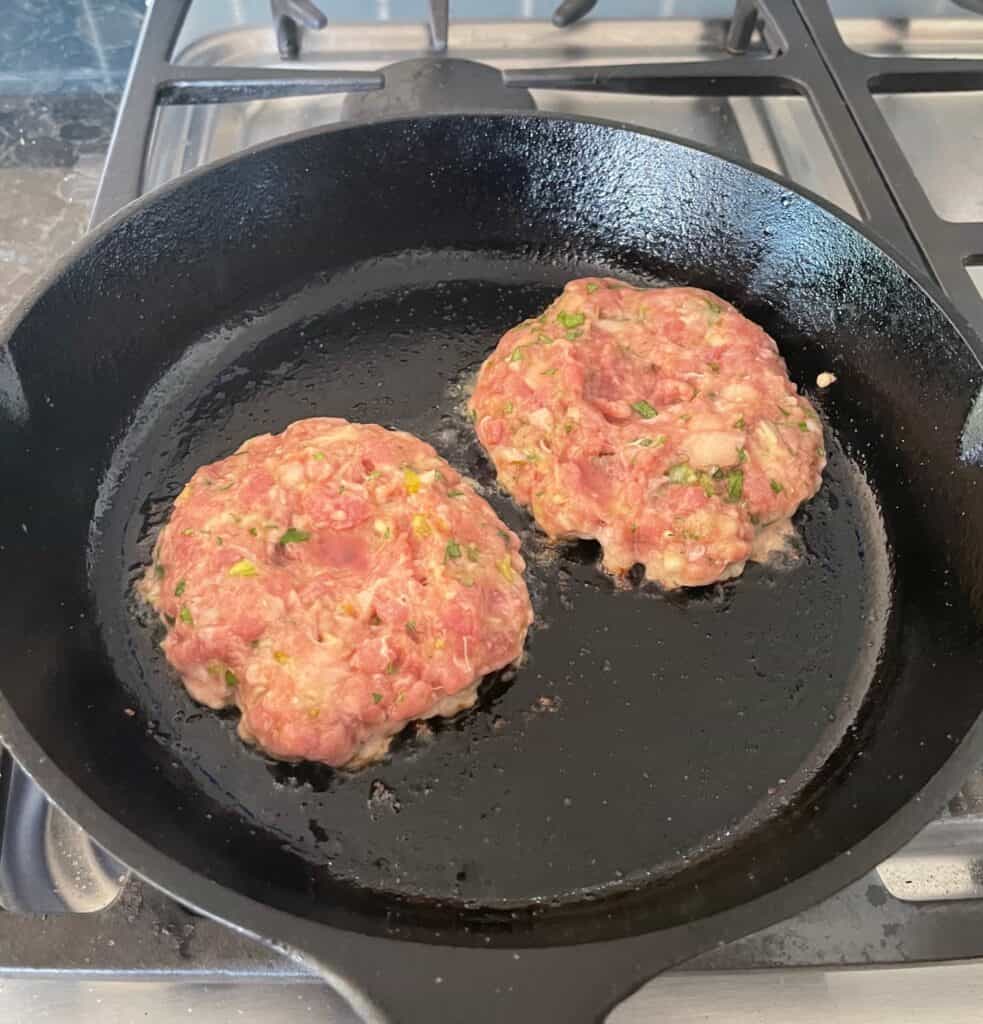 two duck burgers cooking in a cast iron frying pan.