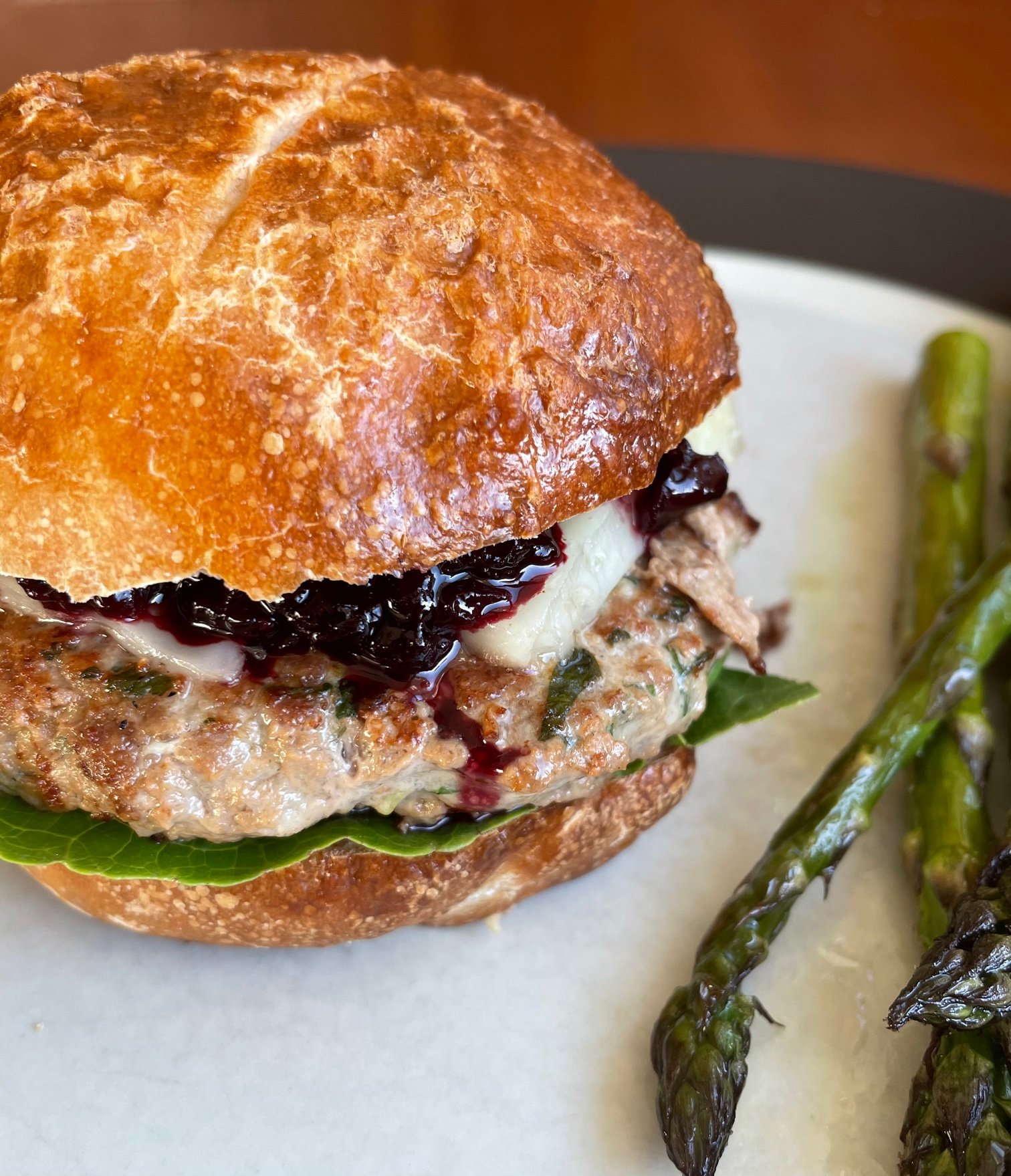 a duck burger on pretzel bun with lettuce, cheese, and cherry ketchup, on a plate with asparagus