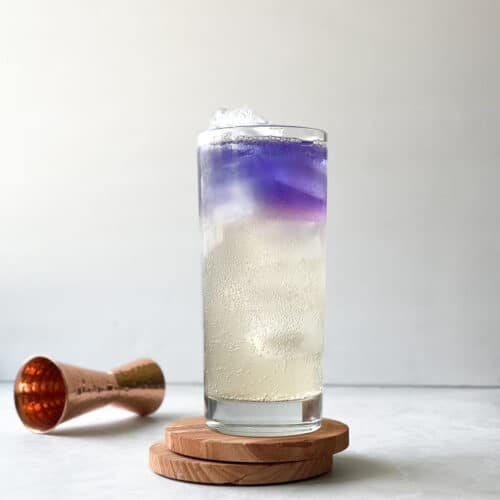 highball glass filled with ice and slightly golden bubbly liquid topped with an inch of deep purple liquid bordered by a bit of pink. The glass sits on two coasters next to a copper jigger.