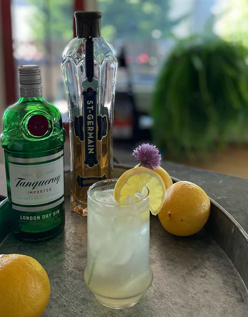 An elderflower Collins garnished with a lemon slice and chive blossom with lemons and liquor bottles