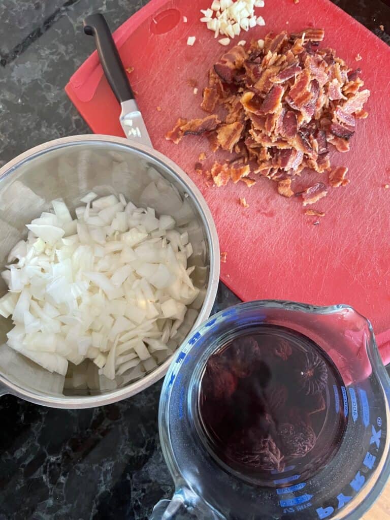 a cutting board with chopped garlic and bacon, onions, and figs soaking in a cup.
