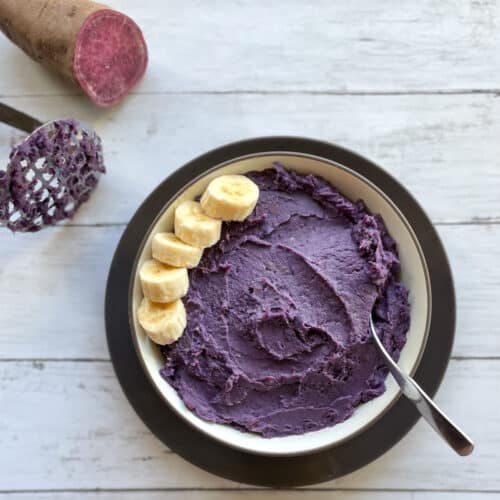 a bowl of purple mashed sweet potatoes garnished with banana next to a potato masher and cut purple sweet potato.