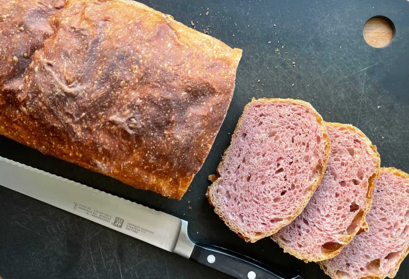 a loaf of pink-tinked purple sweet potato sourdough with three slices and a bread knife.