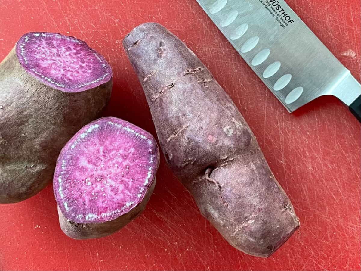 a cutting board with a cut purple sweet potato, a whole potato, and a knife.