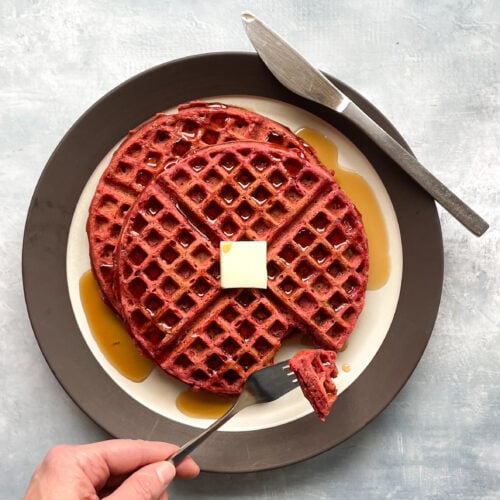 red waffle on a plate with pat of butter and syrup and fork holding a cut piece.