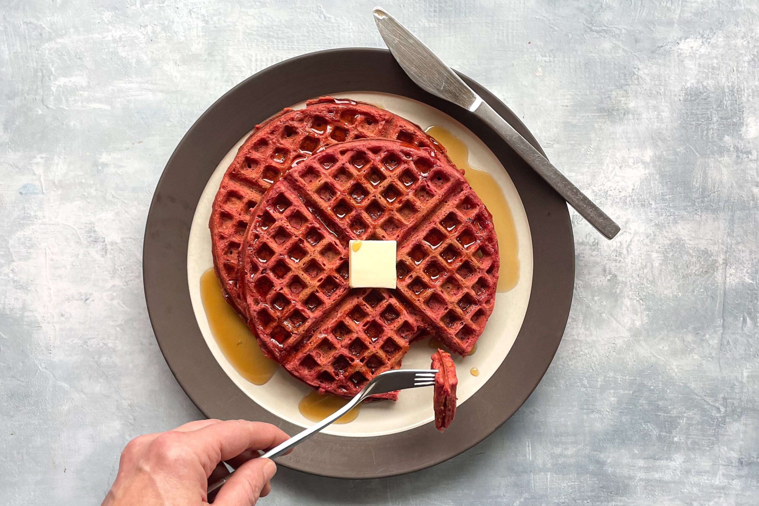 two red waffles on a plate with pat of butter and syrup and fork holding a cut piece.