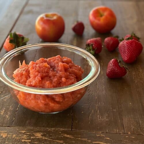 a glass bowl of unsweetened strawberry applesauce surrounded by apples and strawberries.
