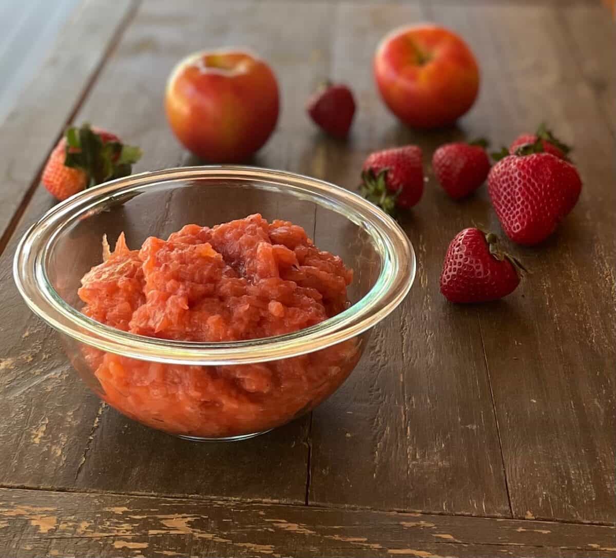 a glass bowl of unsweetened strawberry applesauce surrounded by apples and strawberries.