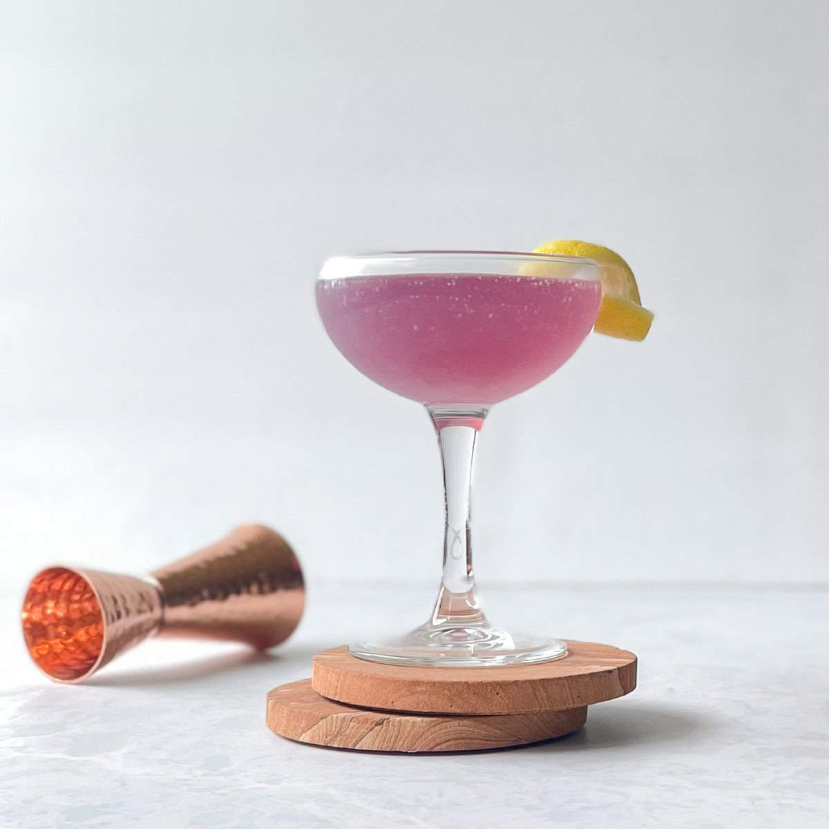 a purple cocktail garnished with a lemon twist in a coupe on two coasters next to a copper jigger.