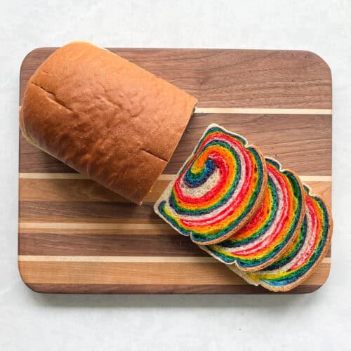 loaf of sandwich bread on a striped wood cutting board. There are three cut slices showing a rainbow swirl.