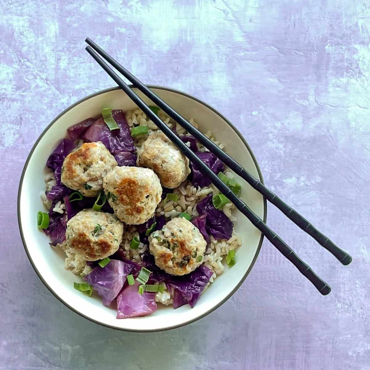 five shrimp and pork meatballs in a rice bowl with purple cabbage. There is a pair of chopsticks resting on the bowl.