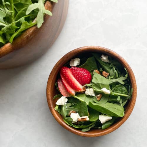 small wooden salad bowl of spinach and arugula with goat cheese and strawberries next to large bowl of greens.