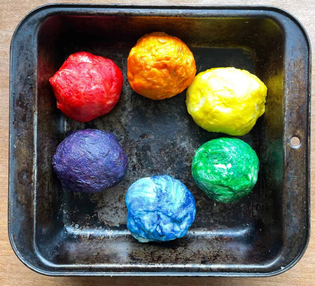 A pan with six rainbow-colored balls of bread dough.