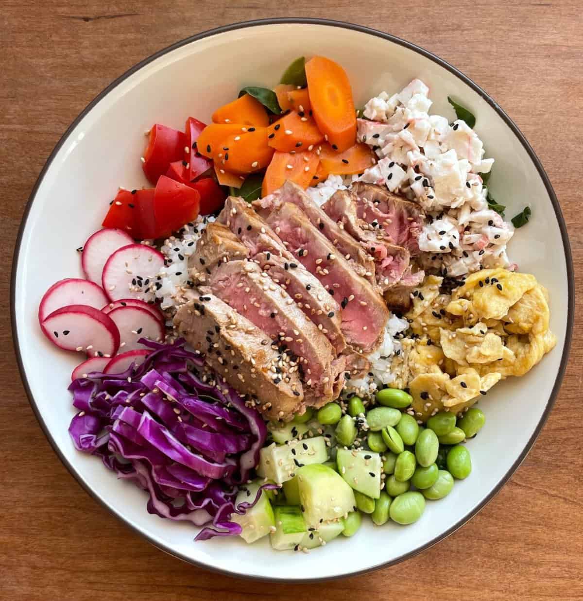 A rainbow poke bowl with sliced seared tuna, sushi crab salad, japanese scrambled eggs, and a rainbow colored array of vegetables.