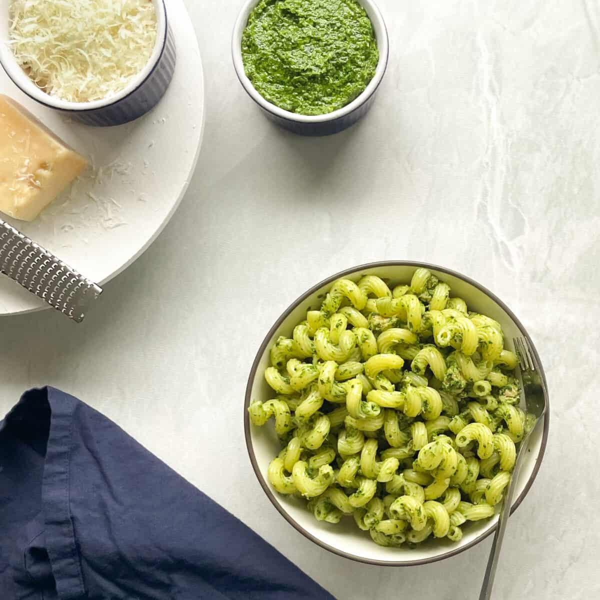 bowls of cavatappi with tuna pesto pasta, arugula spinach pesto, and parmesan with a navy blue napkin.