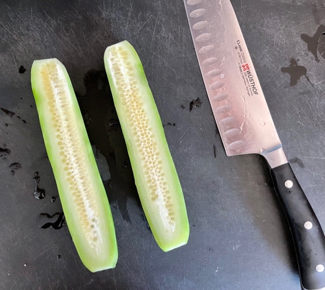 A cutting board with a knife and a cucumber sliced in half.