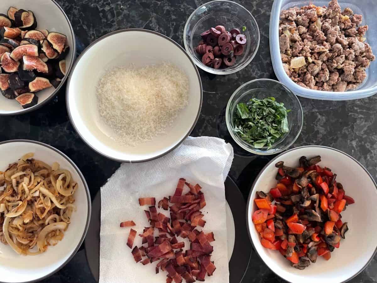 bowls of pizza toppings on a countertop including chopped fresh figs, Parmesan, olives, sausage, basil, roasted red pepper, bacon, and caramelized onions.