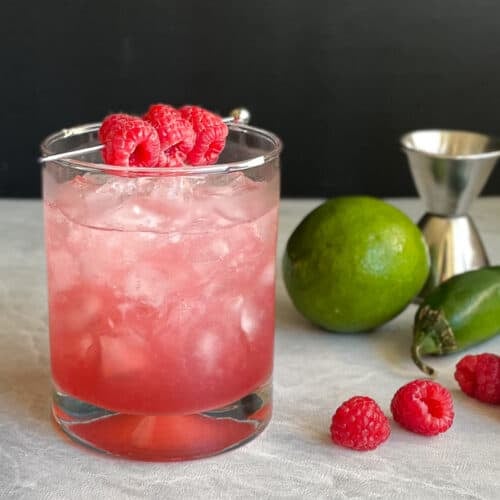 a jalapeno raspberry bramble garnished with three raspberries surrounded by lime, pepper, and berries.