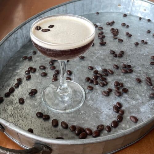 coupe with a dark brown cocktail and three coffee in the foam on a tray surrounded by scattered coffee beans.