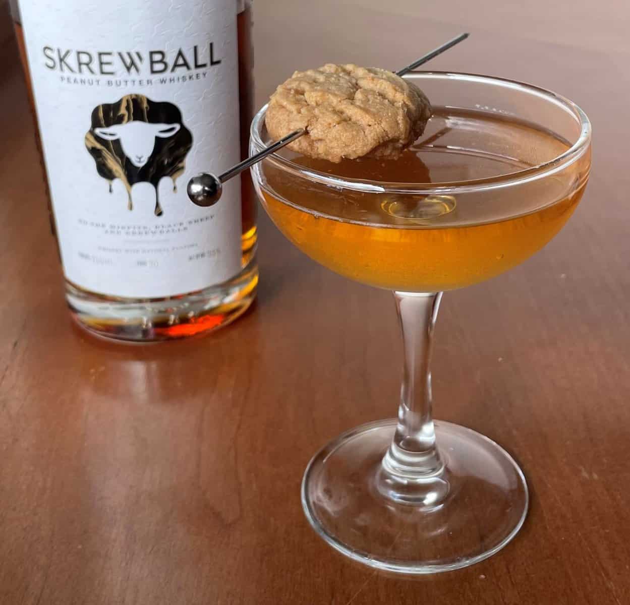 peanut butter manhattan in a coupe garnished with peanut butter cookie next to a bottle of skrewball peanut butter whiskey.