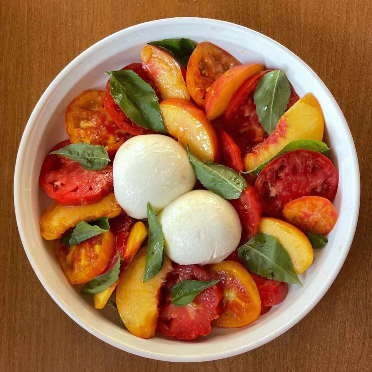 a peach and burrata caprese salad from above with sliced peaches, two different sliced tomatoes, whole basil leaves, and two balls of burrata.