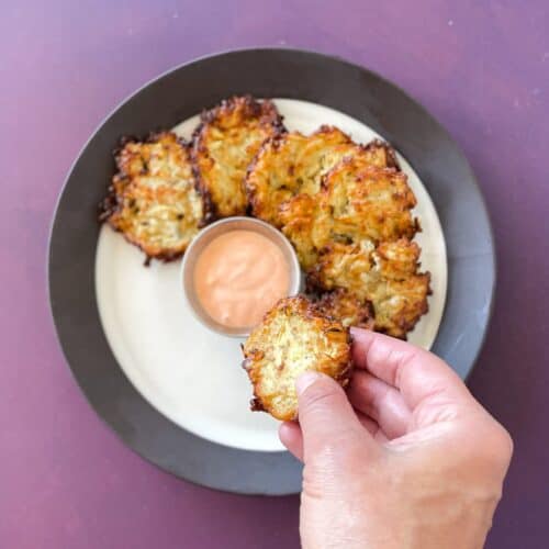 hand holding a fried squash patty over a plate of more summer squash patties and a dip.