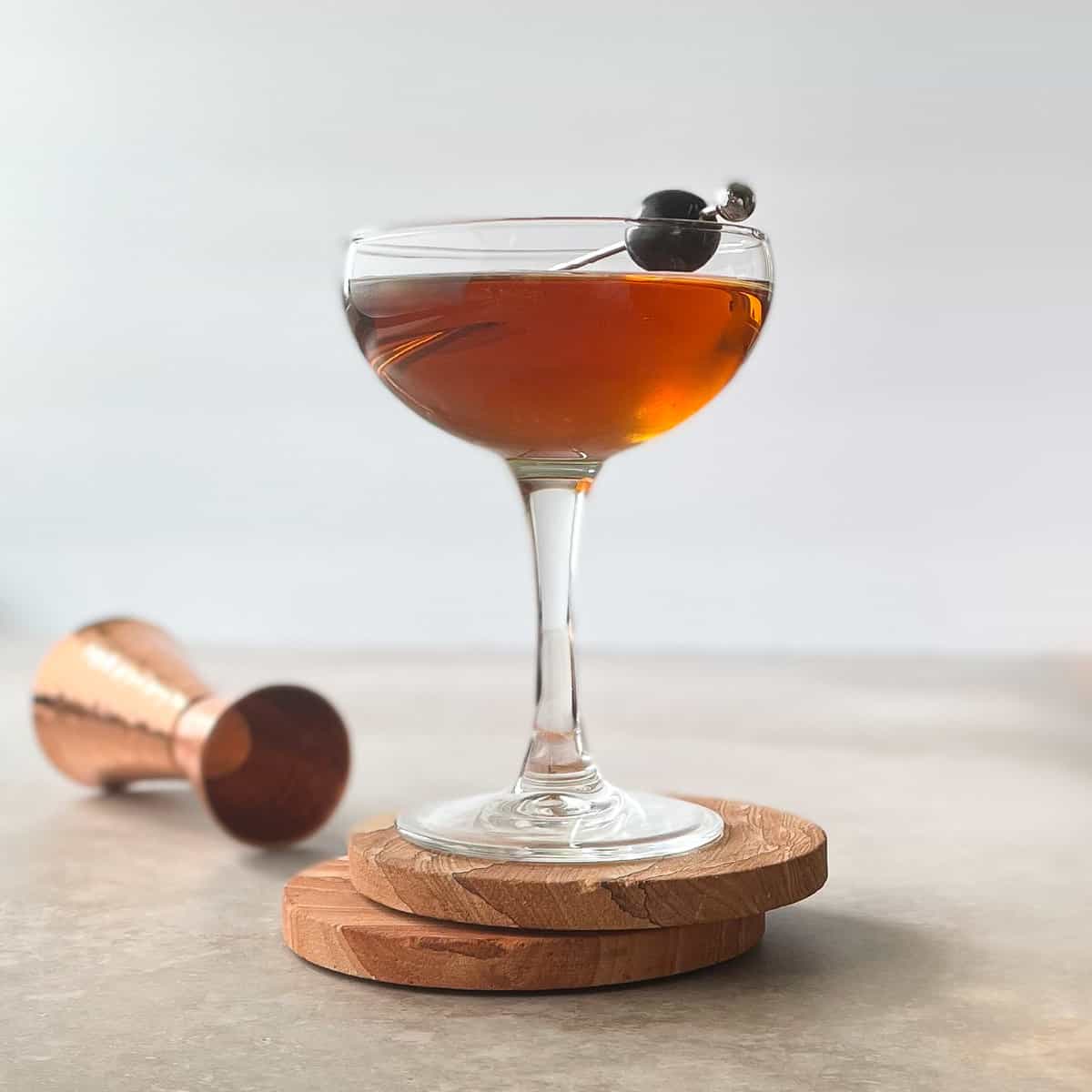 brandy manhattan on coasters garnished with cherry next to an upturned jigger.