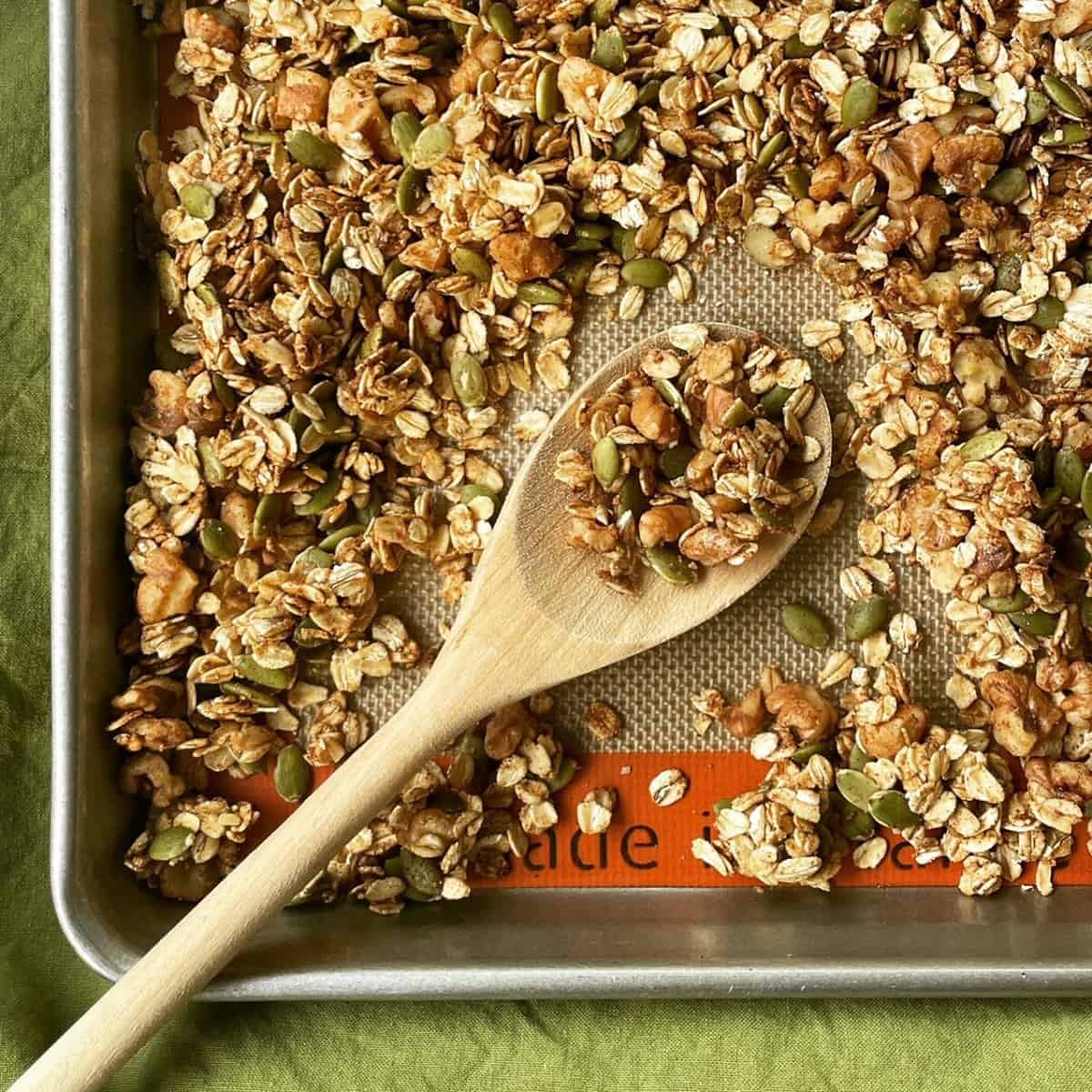 a wooden spoon with a scoopful of granola from a baking sheet filled with homemade granola