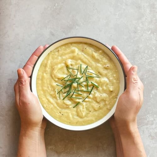 two hands holding a bowl of thick potato soup garnished with chives.