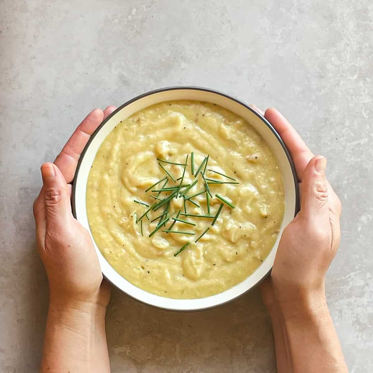 two hands holding a bowl of thick potato soup garnished with chives.