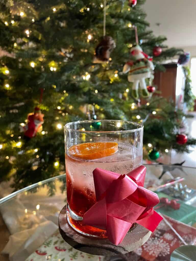 a negroni spritz adorned with a red bow next to a lit christmas tree.