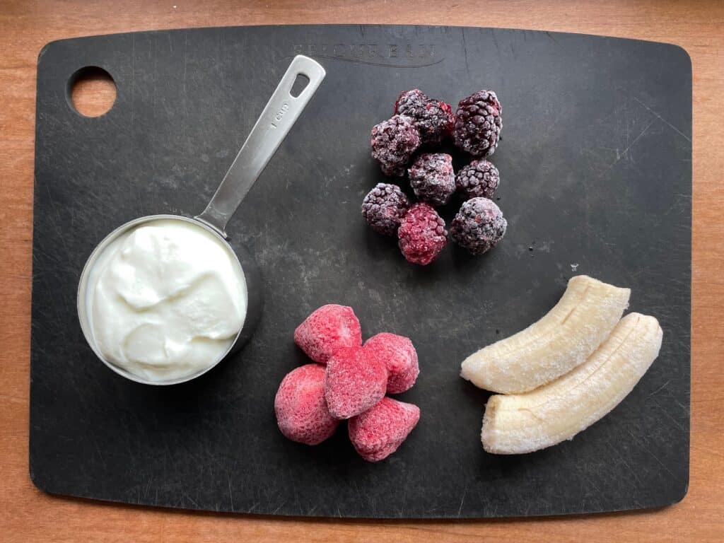 cup of yogurt and frozen blackberries, strawberries, and banana on a cutting board.