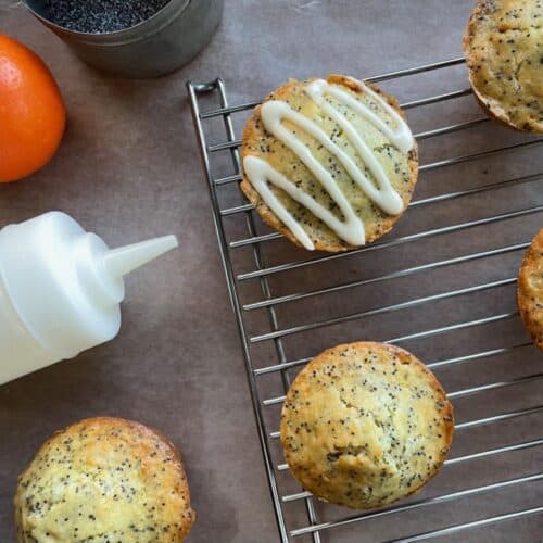 one glazed orange poppy seed muffin and four unglazed muffins on a cooling rack next to an orange, tin of poppy seeds, and glaze in a jar.