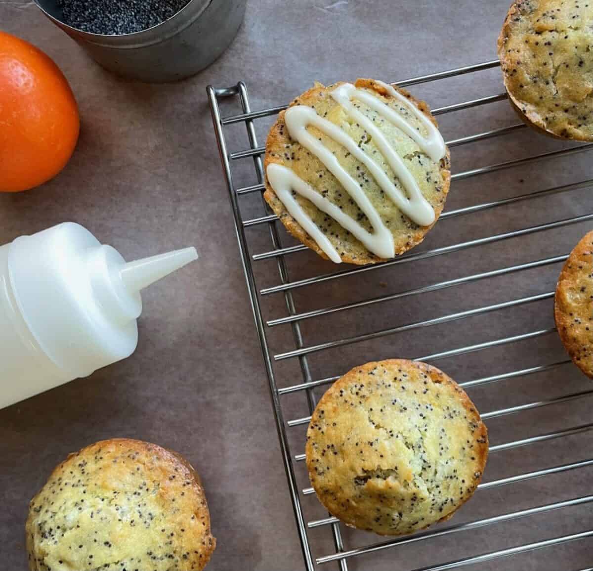 one glazed orange poppy seed muffin and four unglazed muffins on a cooling rack next to an orange, tin of poppy seeds, and glaze in a jar.