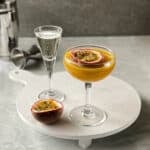 a orange cocktail garnished with a passion fruit half on a white tray with another half and a glass of bubbly.